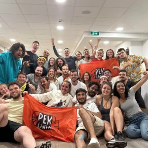 Ensemble des 24 participants et 4 organisateurs de la quatrième aventure et la seconde course qui s’est terminé dans les Alpes. Le groupe se situe à l’intérieur du chalet, tous le monde sourit, certains portent et tendent des drapeaux avec le nom du jeu : Pek Lanta.