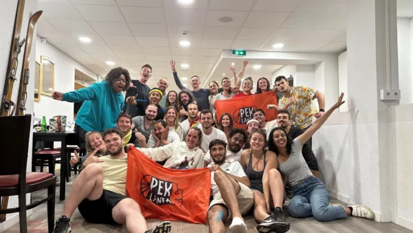 Ensemble des 24 participants et 4 organisateurs de la quatrième aventure et la seconde course qui s’est terminé dans les Alpes. Le groupe se situe à l’intérieur du chalet, tous le monde sourit, certains portent et tendent des drapeaux avec le nom du jeu : Pek Lanta.