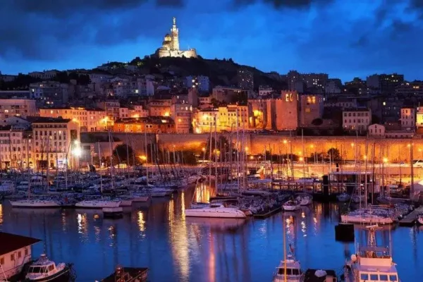 Photo de nuit du Vieux Port de Marseille. Il y a des bateaux, le port et la mer au premier plan, des immeubles au second plan qui recouvre toute la colline et en fond, au sommet de la colline, il y a la Bonne Mère de Marseille, la basilique la plus connue de Marseille, éclairé dans la nuit et dans le ciel noir.
