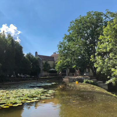 Maison en Bourgogne où s’est déroulé le premier jeu créé par Mauvais joueur en 2021. Un maison avec un moulin à eau sur une rivière, entourée d’arbre en plein Printemps et avec un ciel bleu.